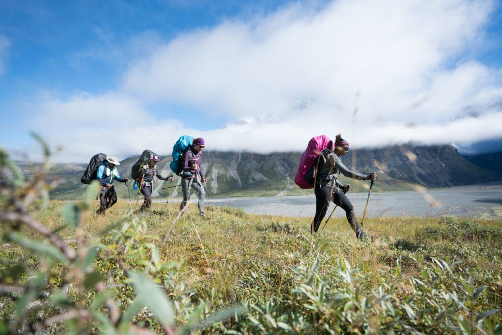 4 people with backpacking packs and trekking poles hiking through a grassland next to a lake