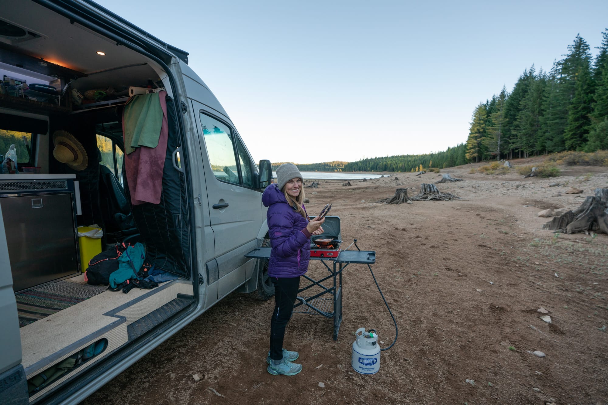 Kristen Bor cooking on a stove in front of her van with water and trees in the distance