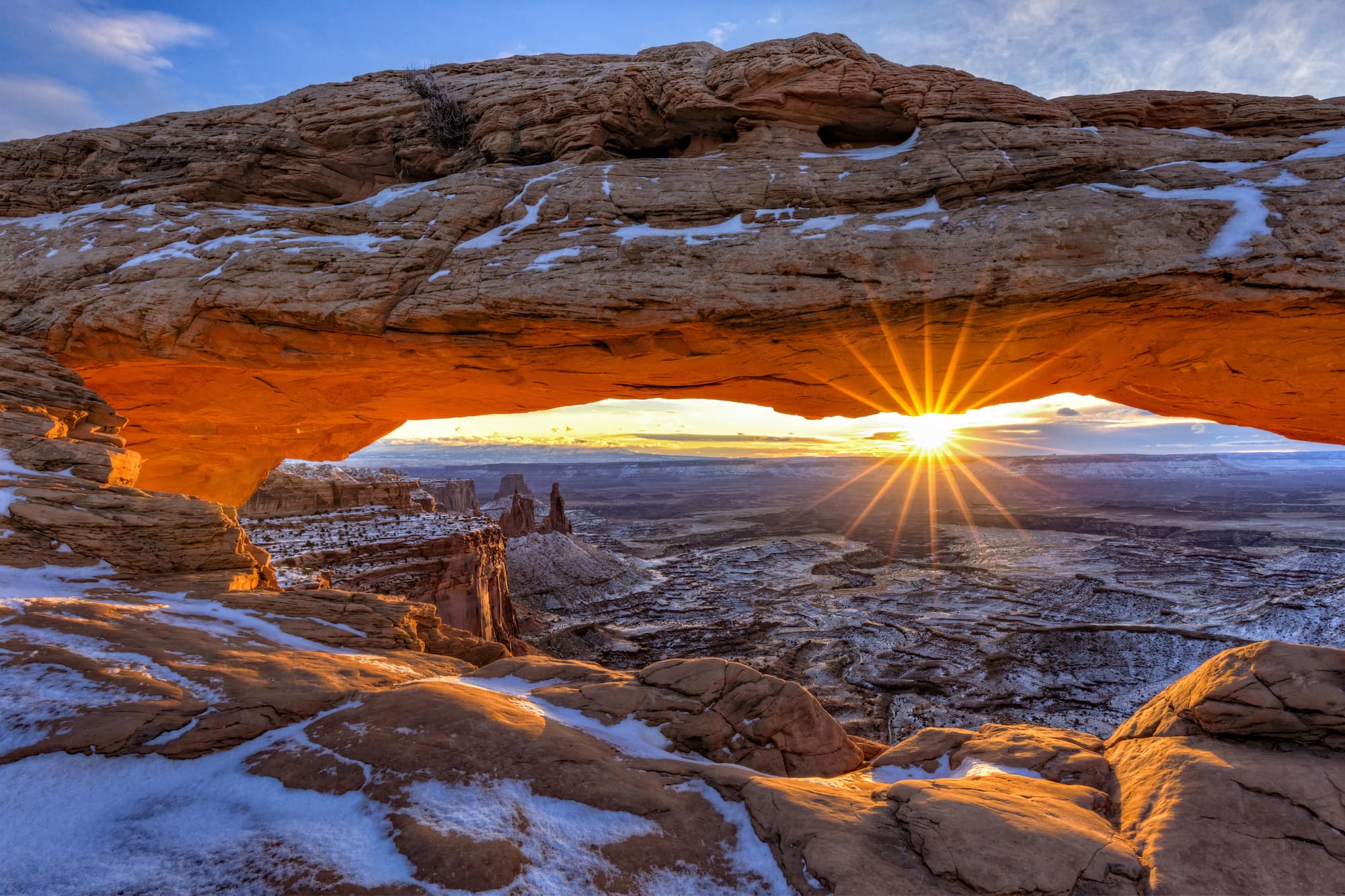 Sun setting behind Mesa Arch in Canyonlands National Park. Red rock landscape is dusted with snow