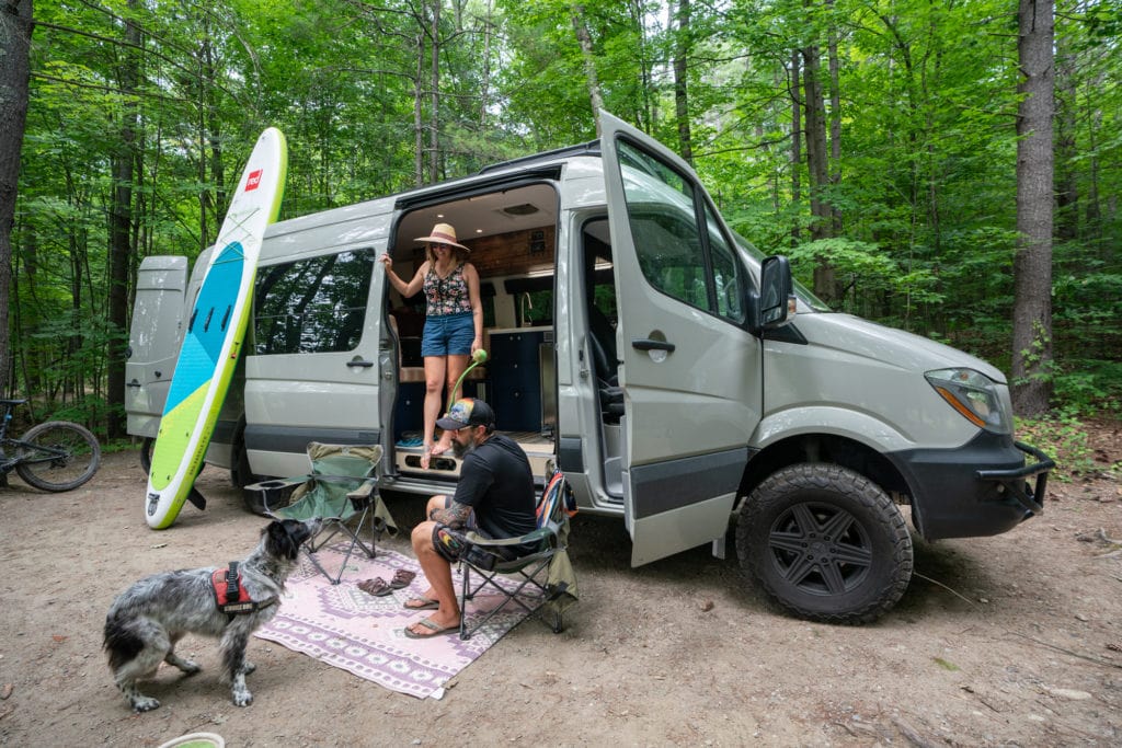 A woman stands in the doorway of a Sprinter camper van and a man sits in a camp chair outside, with a dog and a paddleboard leaned up against the van