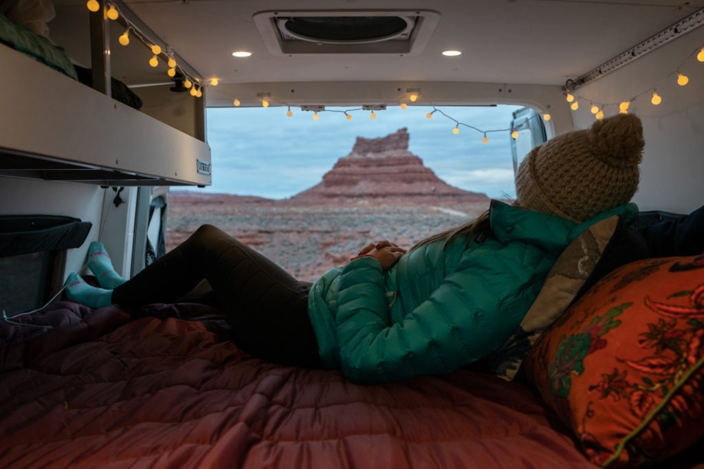 Kristen Bor laying on the bed in a Sprinter van with the back doors open. She is laying on a Rumpl blanket and looking out at a view of a red rock formation.