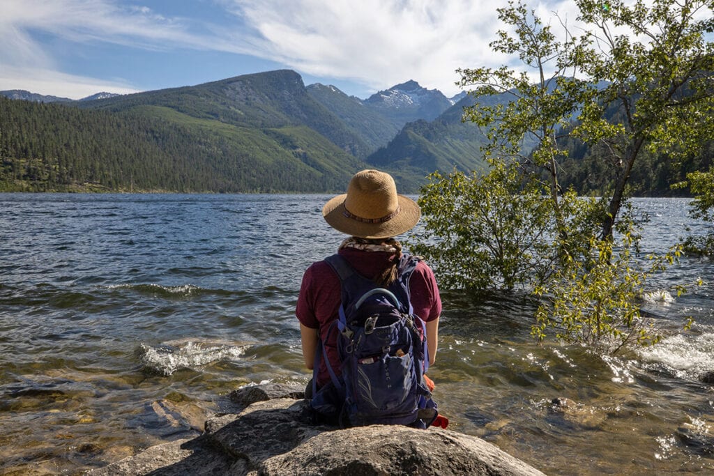 A woman sits on a rock facing a lake. She is wearing a hiking sun hat and day pack