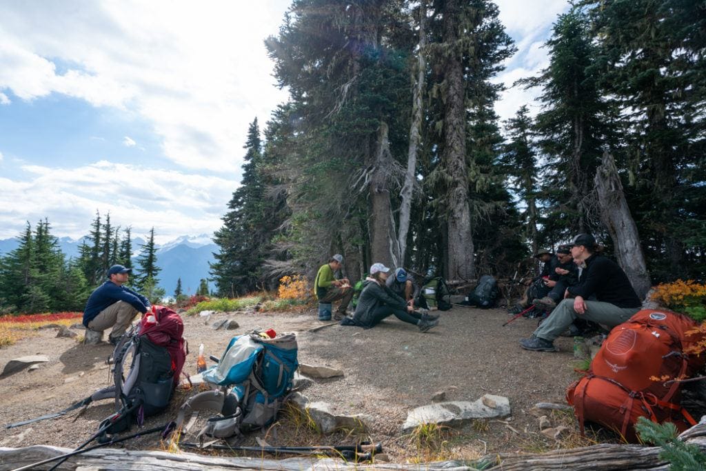 A group of backpackers sit taking a break on the trail. Several backpacking packs are sitting on the ground nearby.