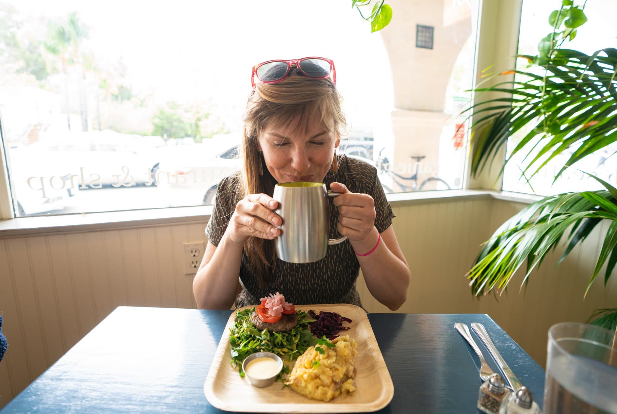 Woman sitting at counter sipping from stainless steel mug with plate of supplies in front of her