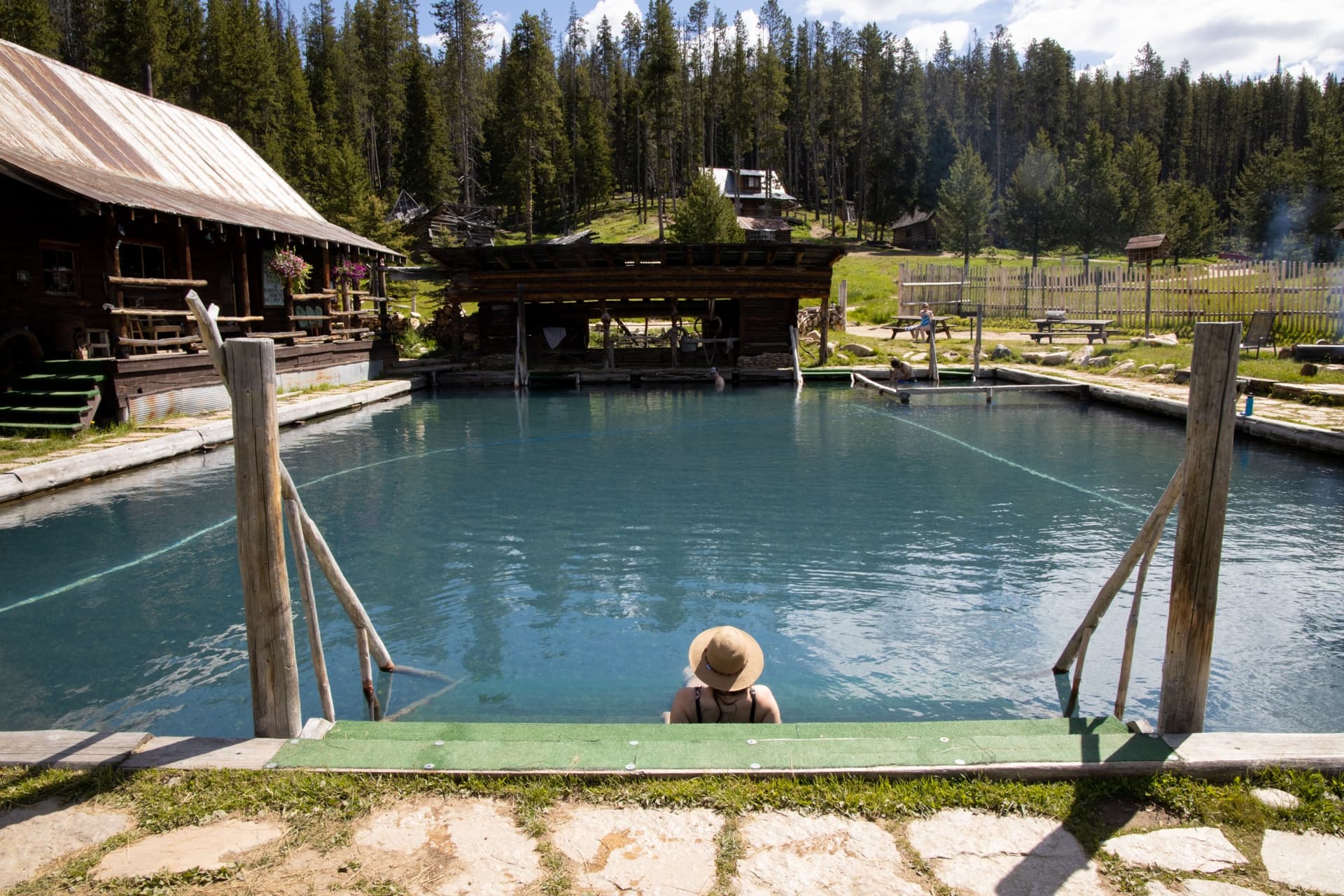 Woman soaking in Burgdorf hot spring pool in Idaho surrounded by pine trees and historic wooden buildings