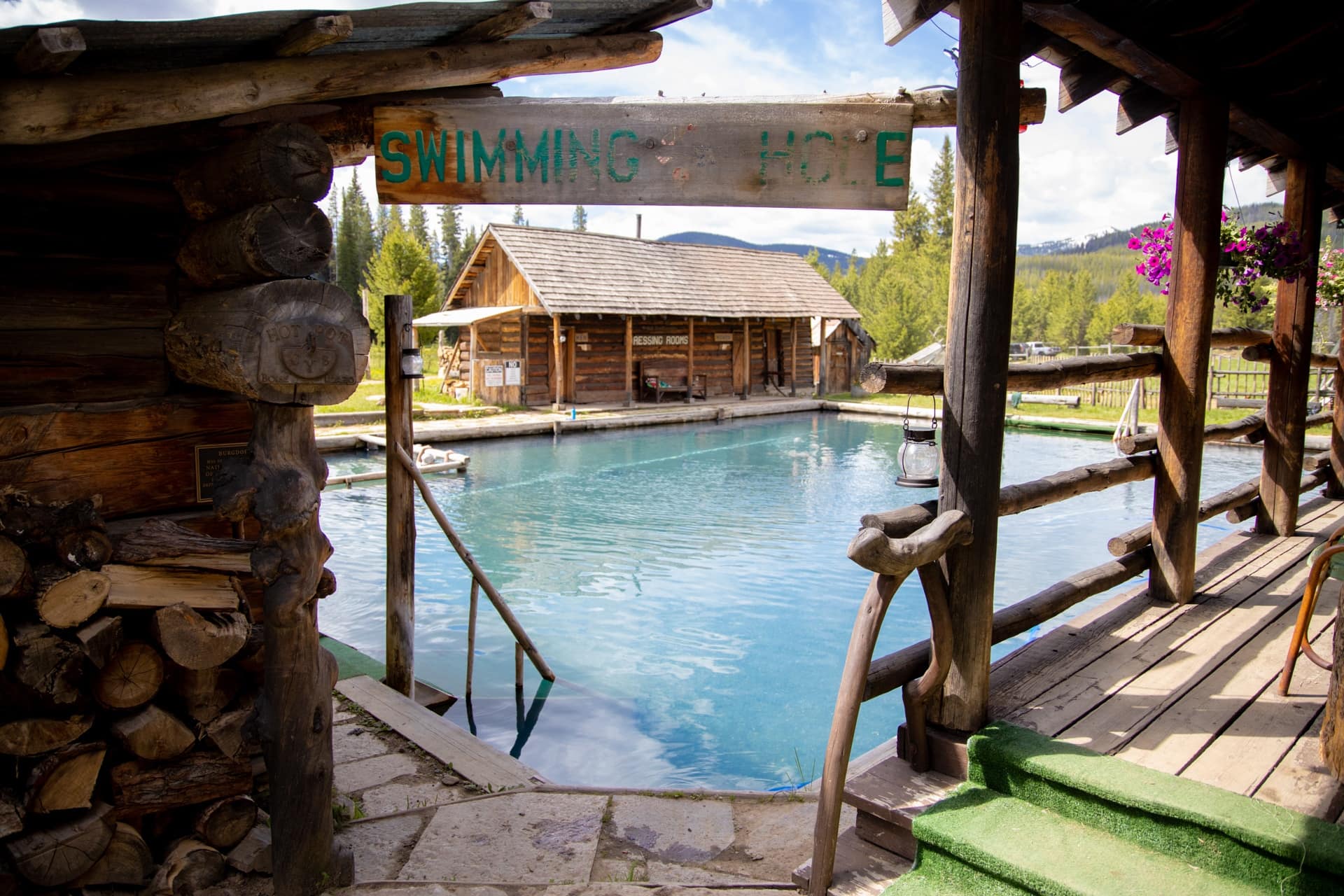 A large hot spring pool at Burgdorf hot springs resort in Idaho. Log cabin buildings surround the pool and sign saying "swimming hole" hung over entrance into pool