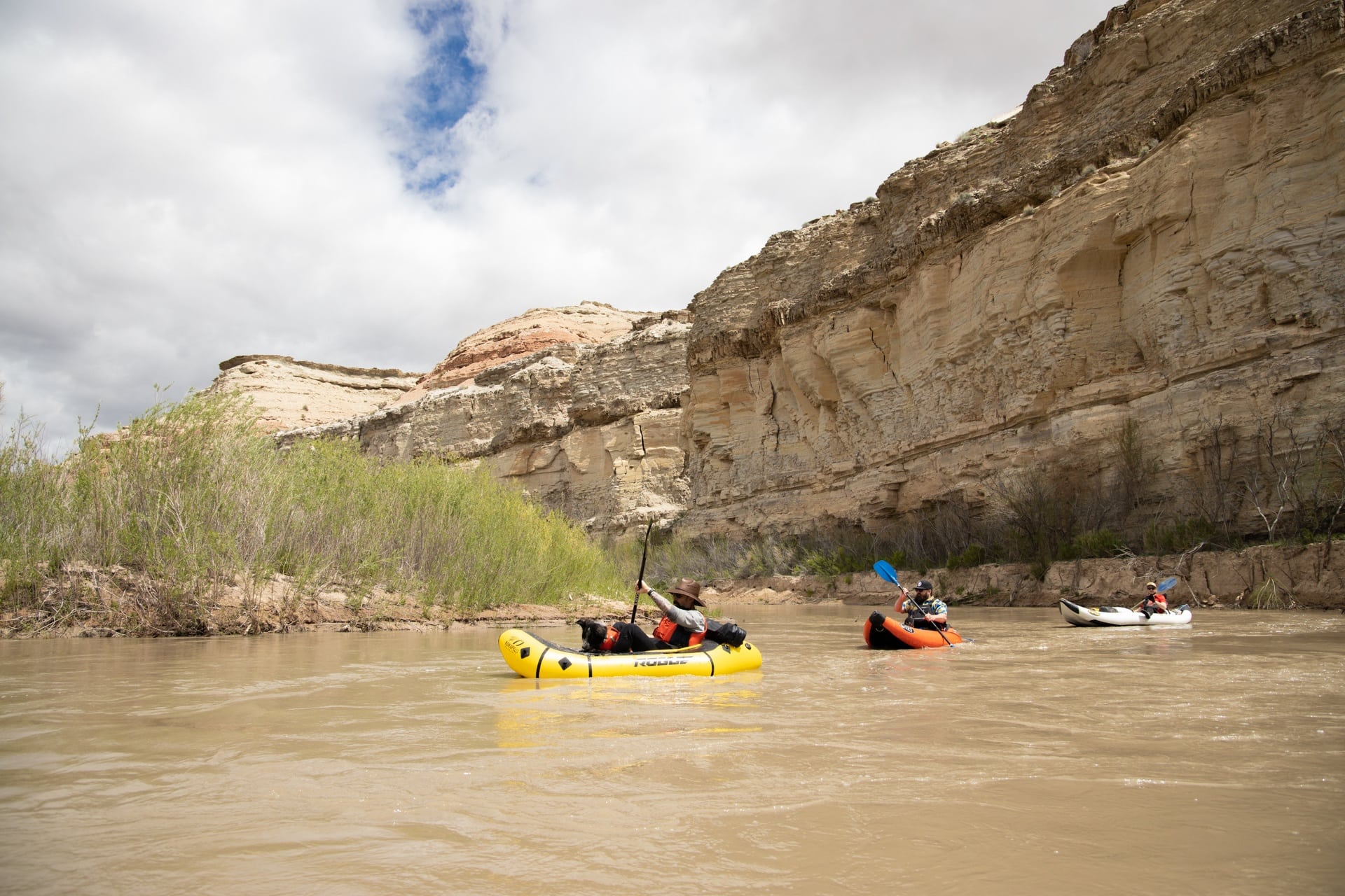 Three people in packrafts floating down the San Rafael River in Utah with steep rock cliffs forming one edge of the river