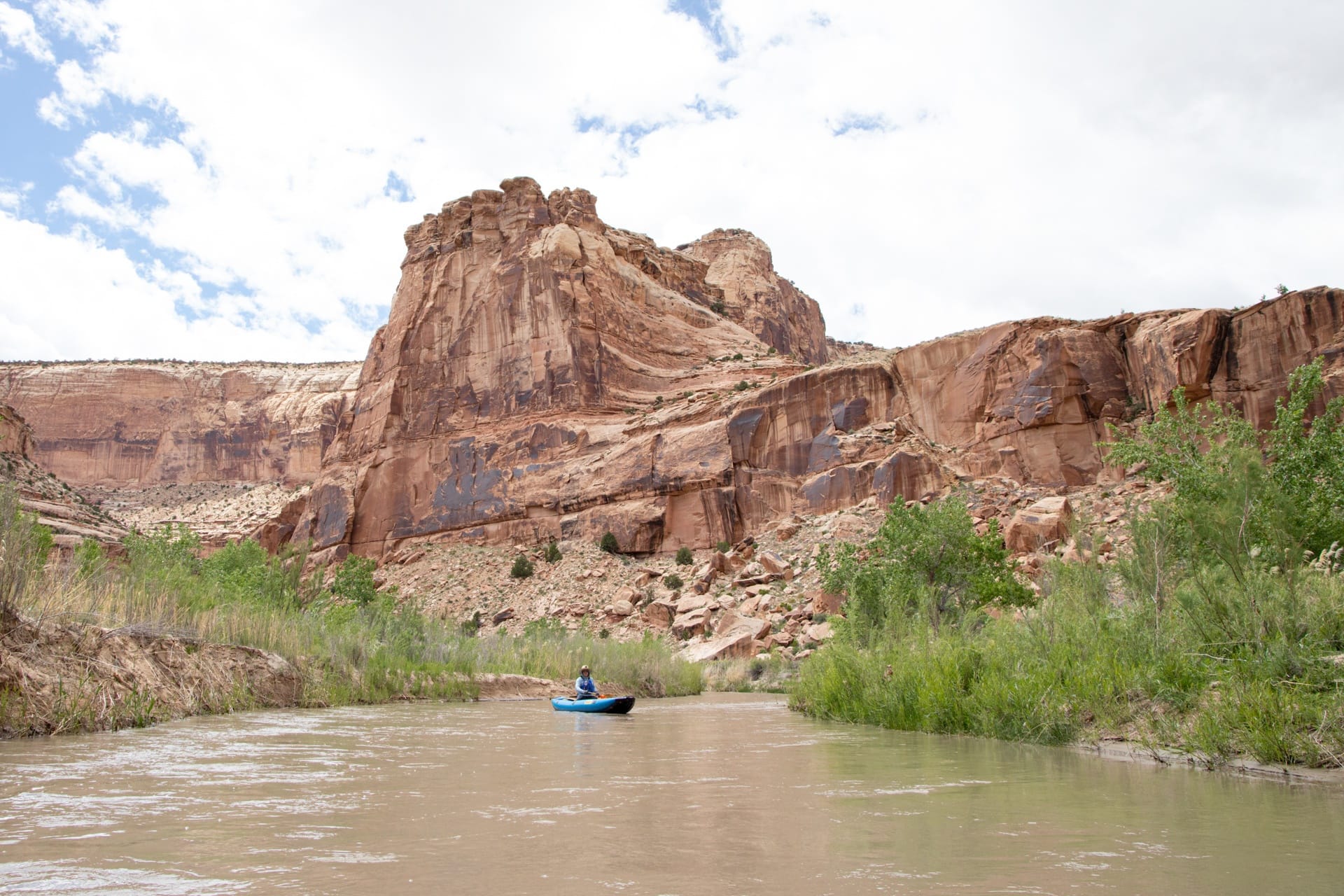 Person in pack raft on San Rafael River in Utah with tall red rock bluffs behind her and green vegetation lining river banks