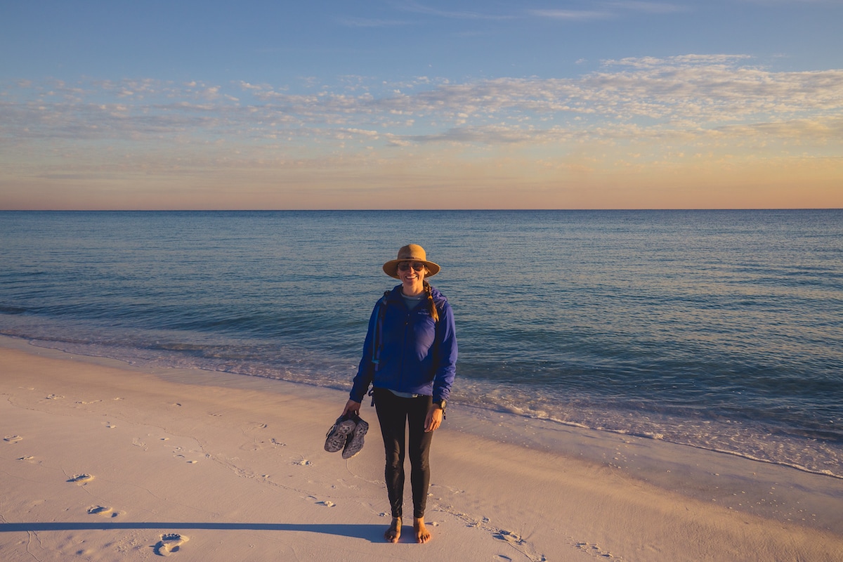 Kristen Bor smiling on a sandy beach with the ocean and sunset behind her while wearing the Wallaroo Sedona Hat and holding a pair of shoes