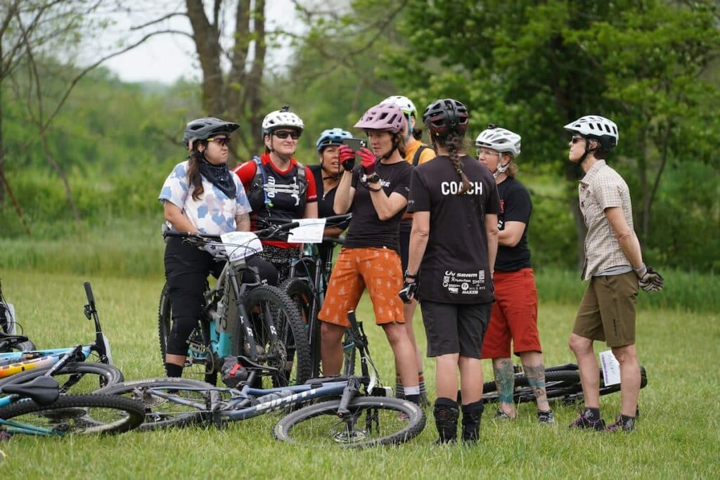 Group of female mountain bikers crowded around coach to watch video on a phone during a coaching clinic