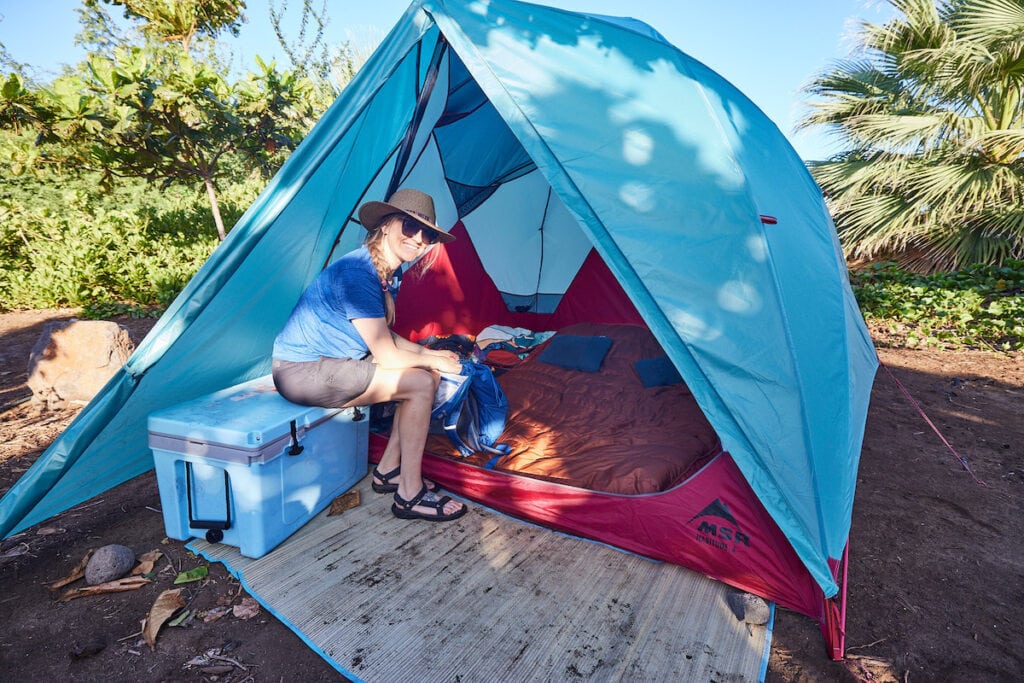 A woman sits on top of an RTIC cooler outside of a MSR Habitude Tent while car camping