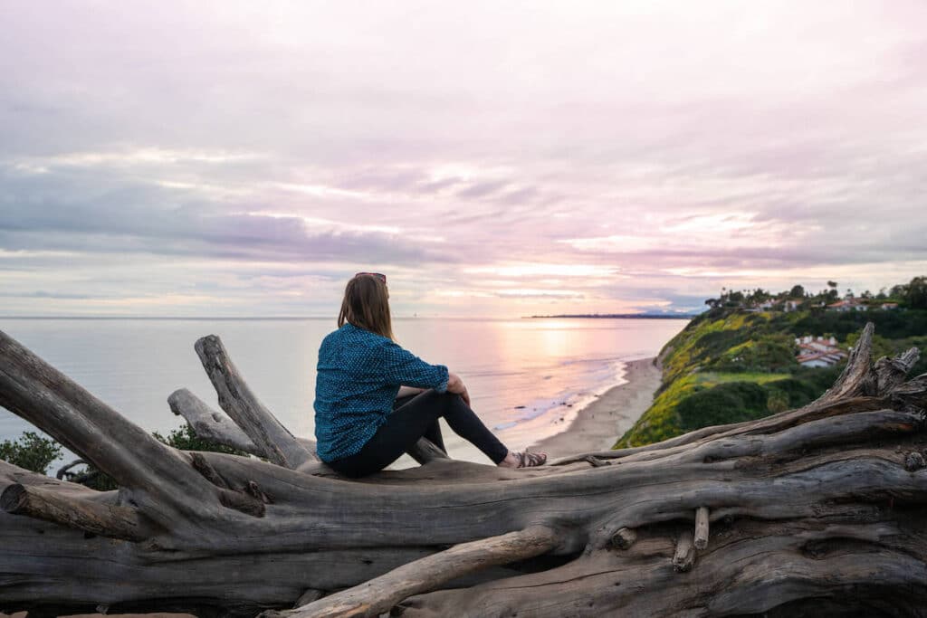 Woman sitting on large piece of driftwood watching sunset off the coast of California 