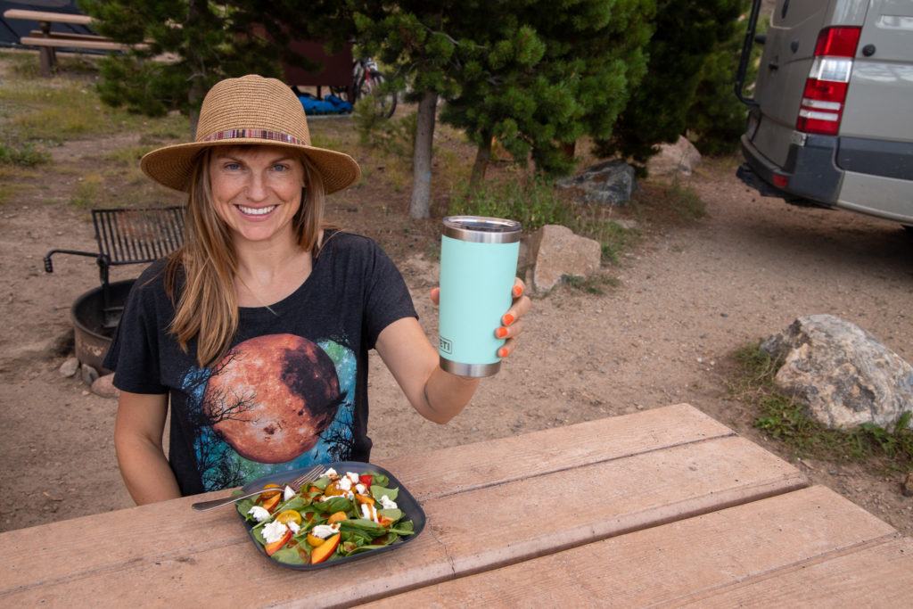 Kristen sitting at picnic table with plate of food in front of her and raised insulated mug in one hand