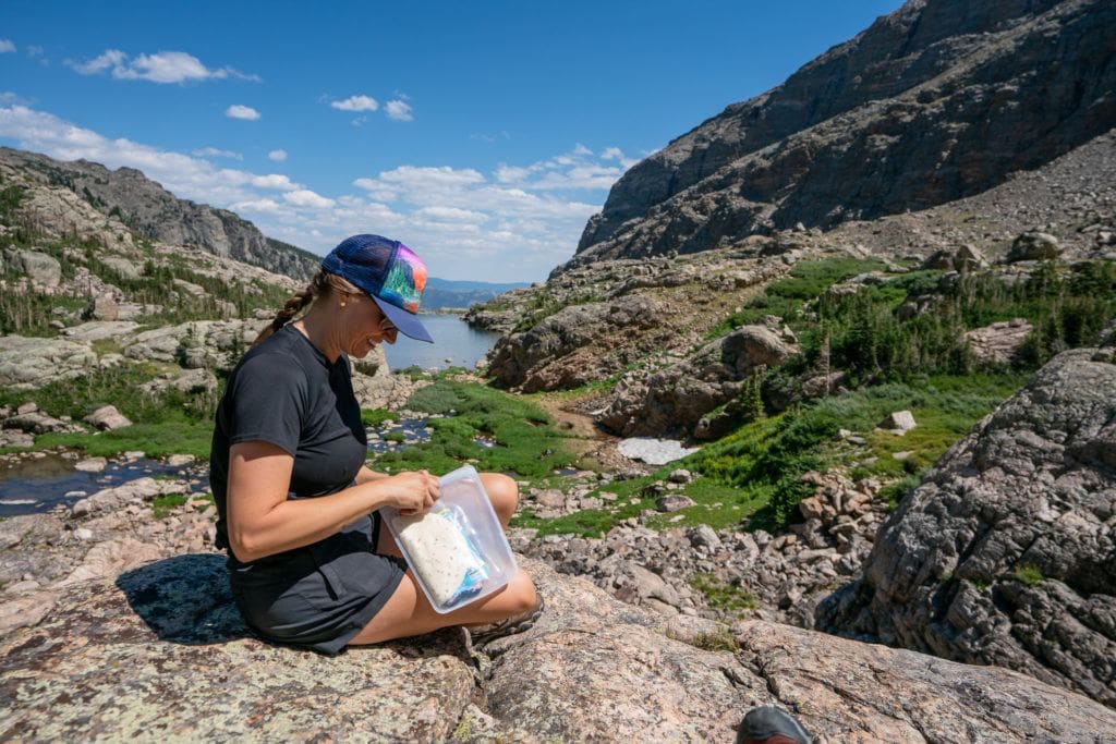 Woman sitting on rock on hike eating snacks out of a reusable Stasher Bag