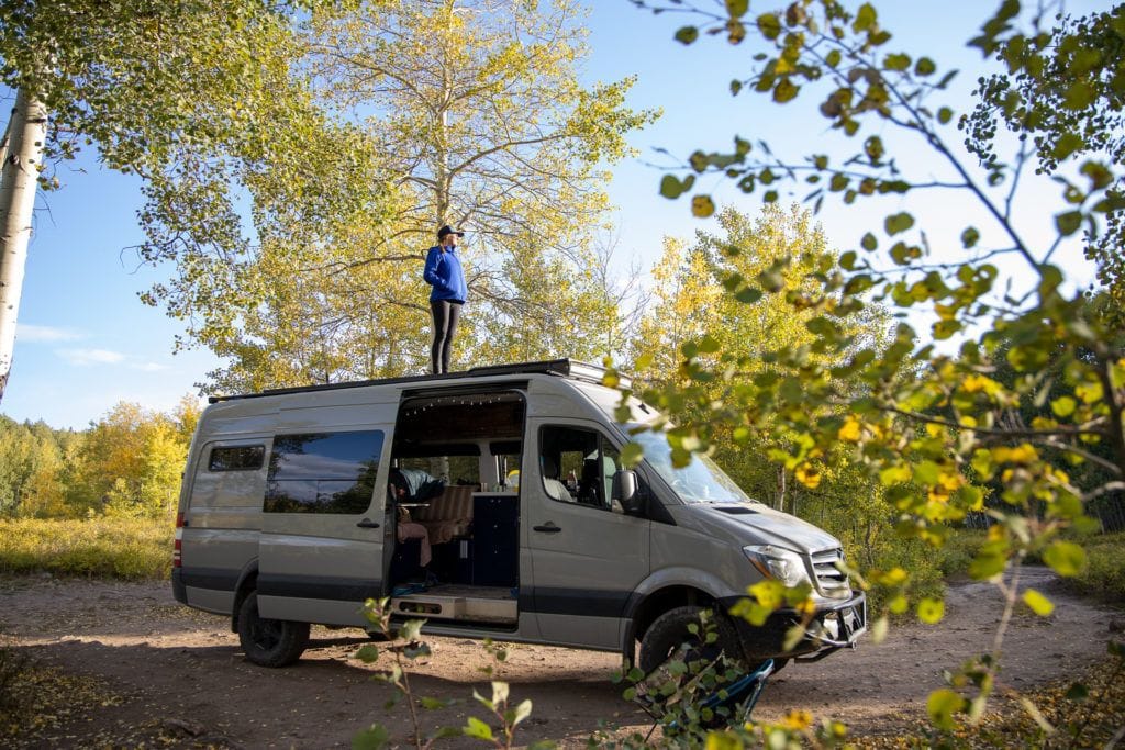 Kristen standing on top of her converted Sprinter camper van at 
a campground surrounded by trees