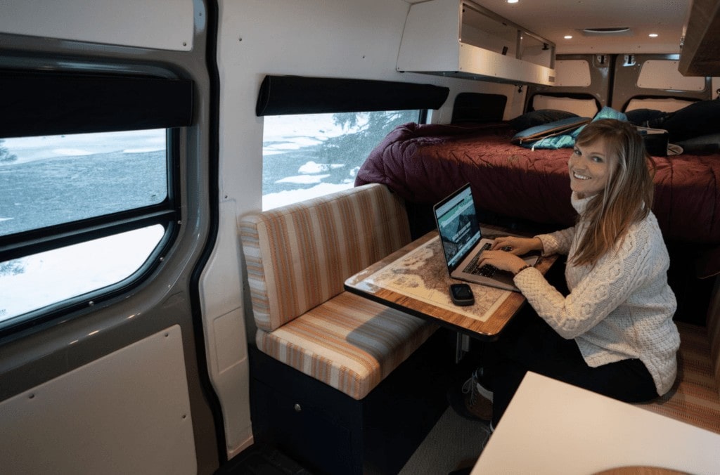 A woman sits on her laptop smiling at the camera. She is sitting at a desk in her camper van