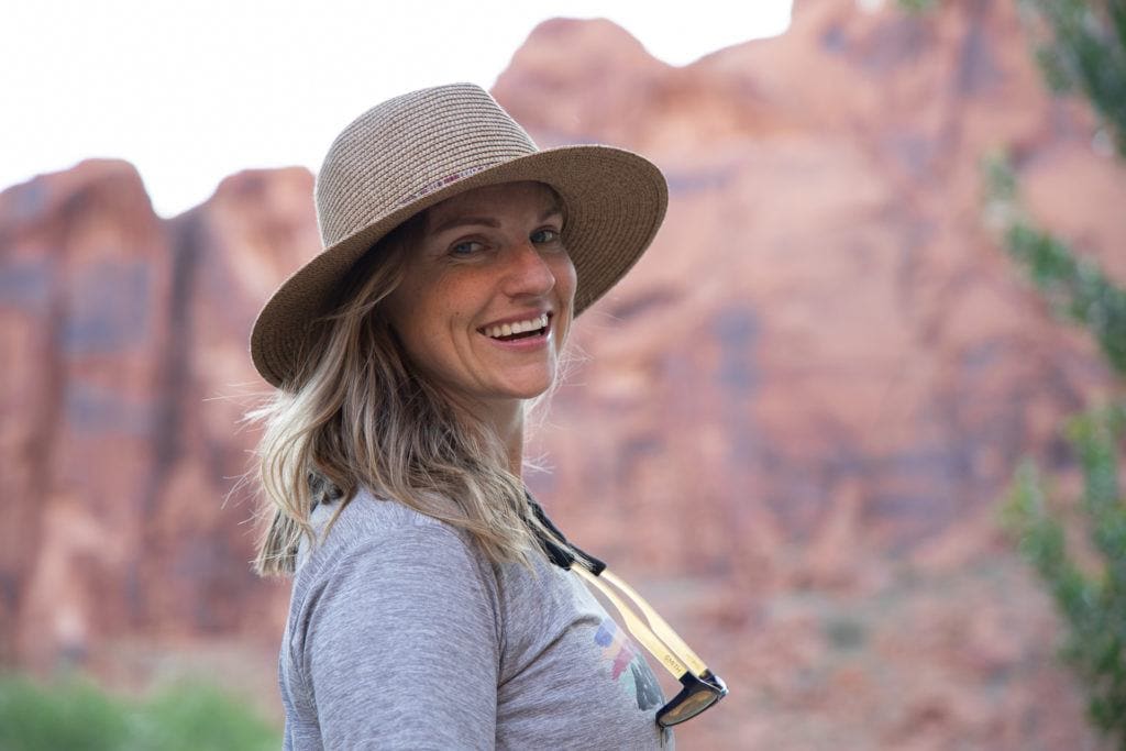 Portrait shot of woman smiling at camera wearing wide brimmed sun hat and sunglasses around her neck