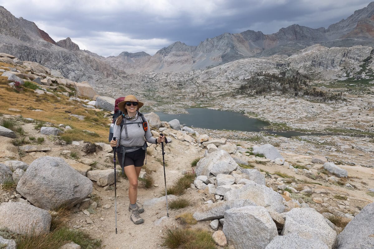 Woman hiking on trail in high alpine terrain carrying backpacking gear and holding trekking poles
