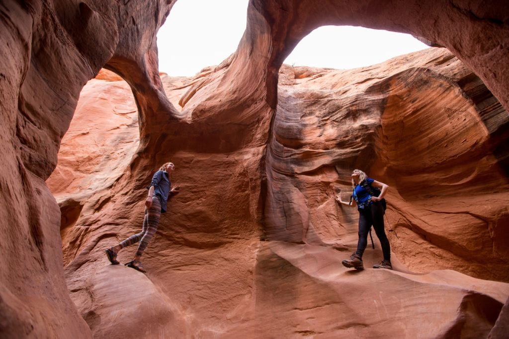Two women exploring red rock cave with three sky holes in Utah