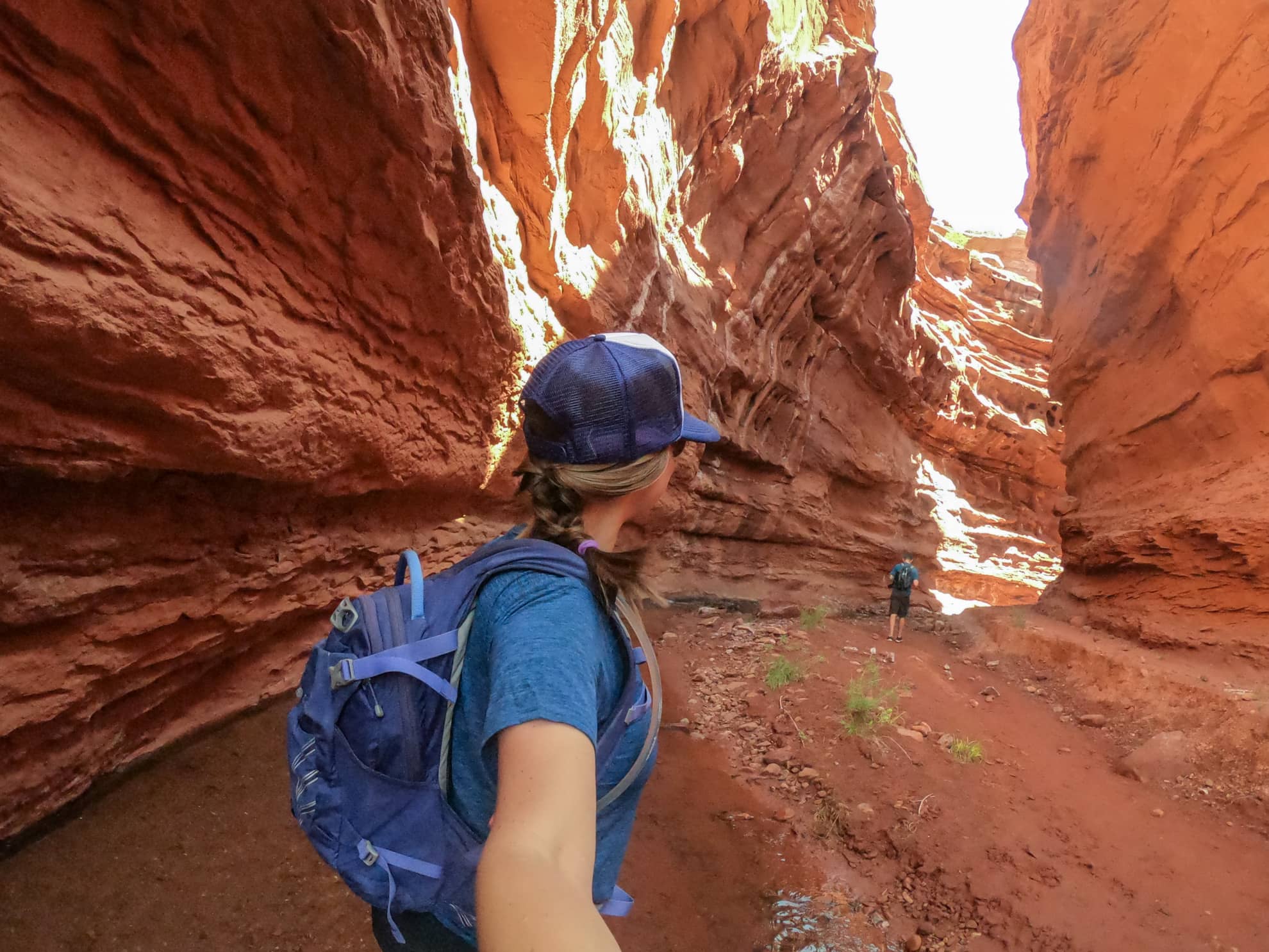 Hiker taking a selfie while trekking through narrow slot canyon with tall cliff walls on either side and standing water 