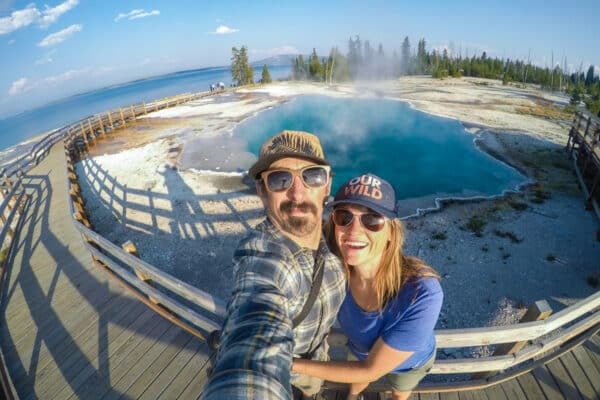 Kristen Bor and her partner taking a selfie next to a blue geyser in Yellowstone's West Thumb Geyser Basin with Yellowstone Lake in the Background