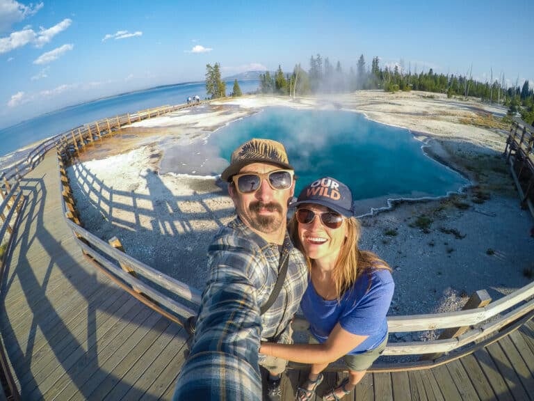 Kristen Bor and her partner taking a selfie next to a blue geyser in Yellowstone's West Thumb Geyser Basin with Yellowstone Lake in the Background