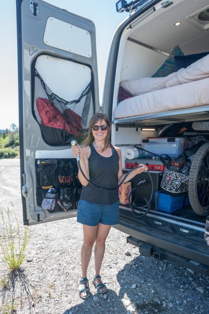 woman smiling holding a shower head with the rear doors of a Sprinter van open showing off her van outdoor shower