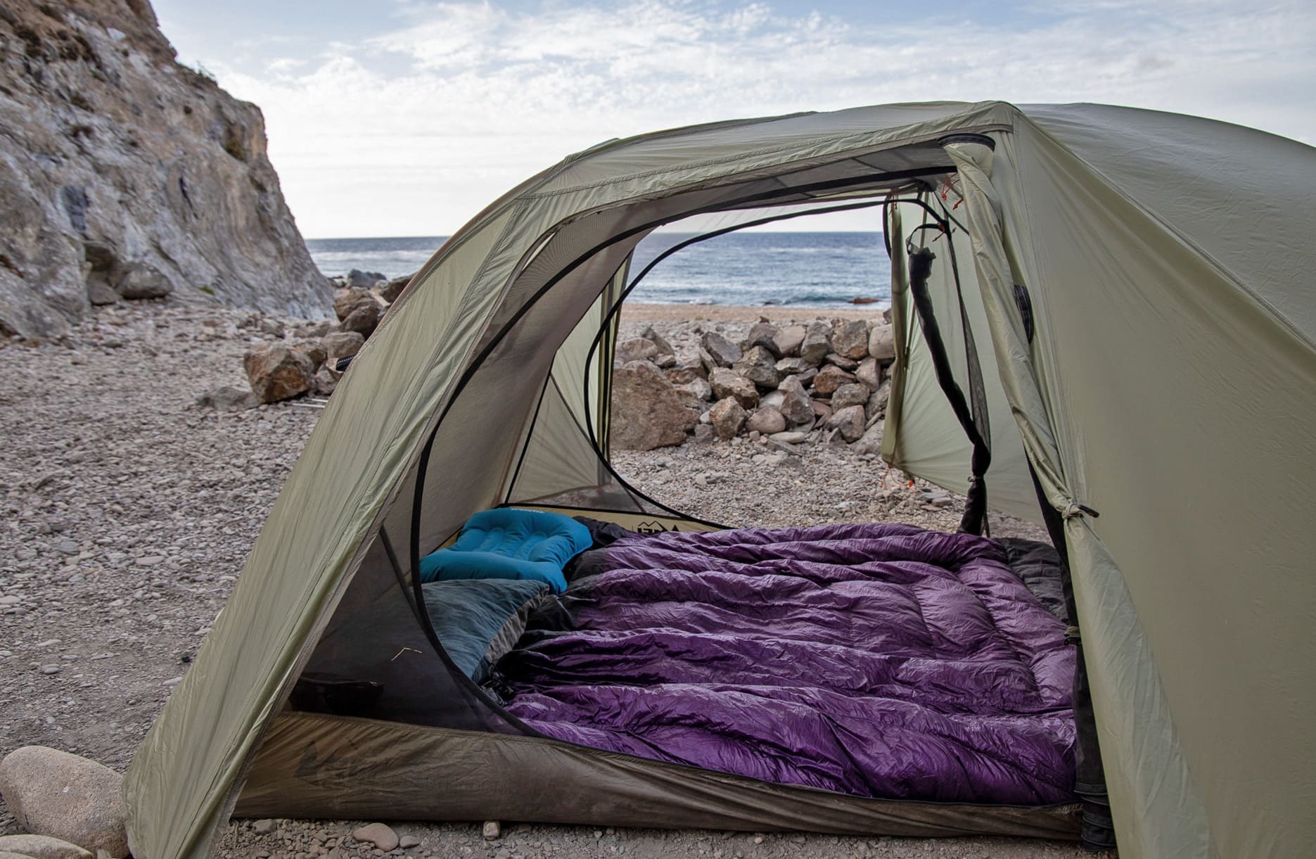 a backpacking tent set up next to the ocean with a purple 2-person backpacking quilt laid out