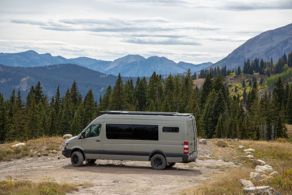 Sprinter van parked in dirt circle surrounded by pine trees with Colorado mountain range in distance. 