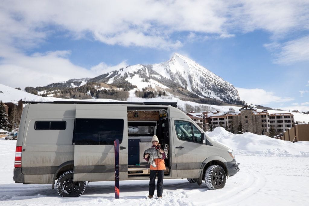 Kristen in front of sprinter van holding dog in a winter ski destination with snowy mountain in background.