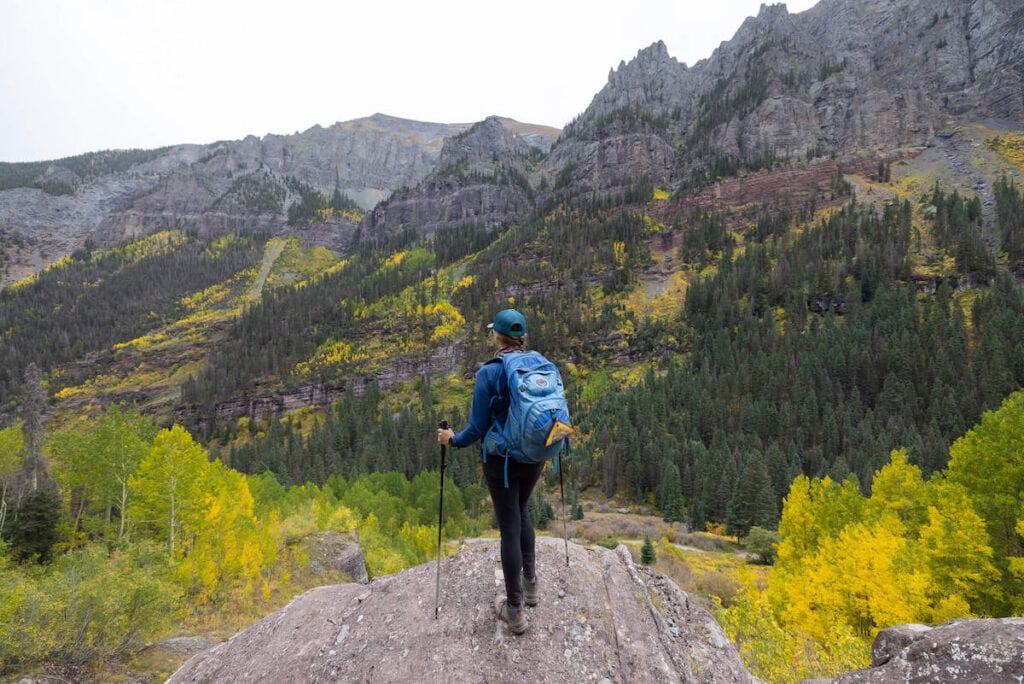 Kristen standing on rock ledge on hike in Colorado looking out over mountain range wearing blue Osprey backpack and holding hiking poles