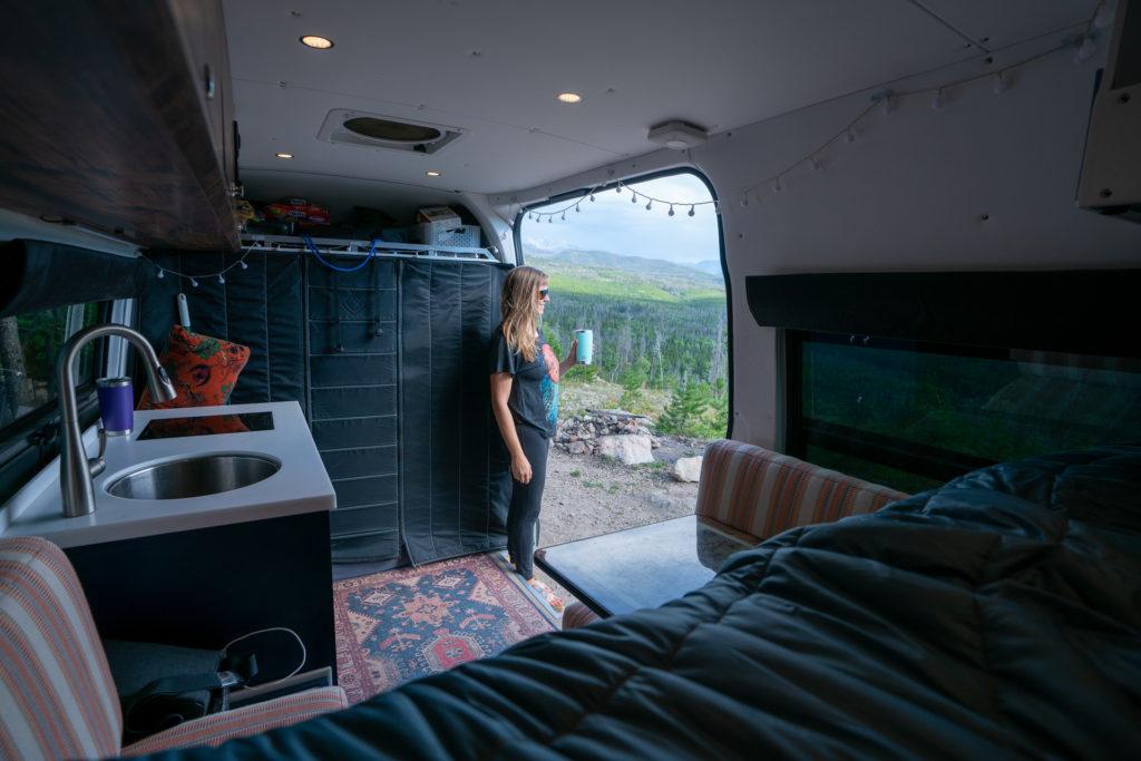 a woman holding a blue tumbler stands in the doorway smiling while looking outside of her camper van