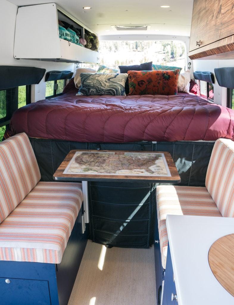 the interior of a campervan with a full size bed in the back with table with two seating chairs on either sid