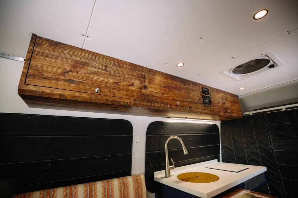 Interior of converted camper van with wooden cabinets hanging from ceiling and kitchen and sink area below. Windows covered with blackout curtains.