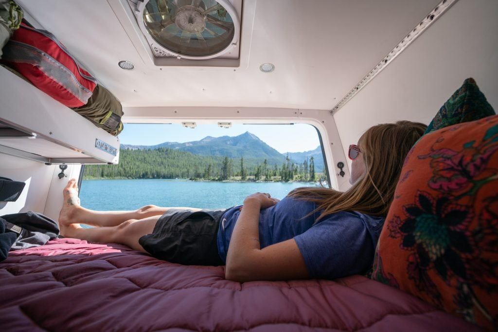 Kristen lying on bed in converted Sprinter Van with back doors open looking out onto scenic lake view with mountains