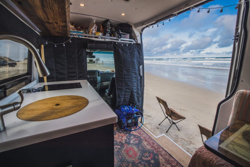 Converted Sprinter van kitchen area with sliding door open to white sand beach