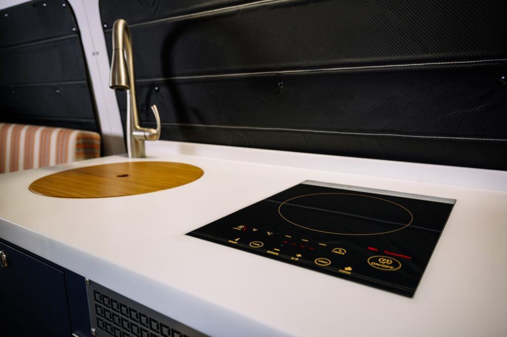 Induction stove inlaid into white counter in converted camper van next to sink and faucet