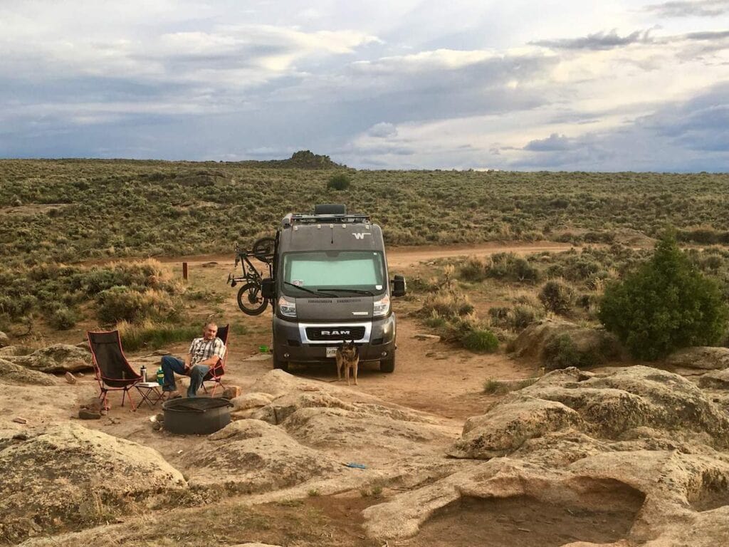 Black van facing camera parked in high desert dispersed campsite. Man sitting in camp chairs in front of fire ring and dog standing in front of van 