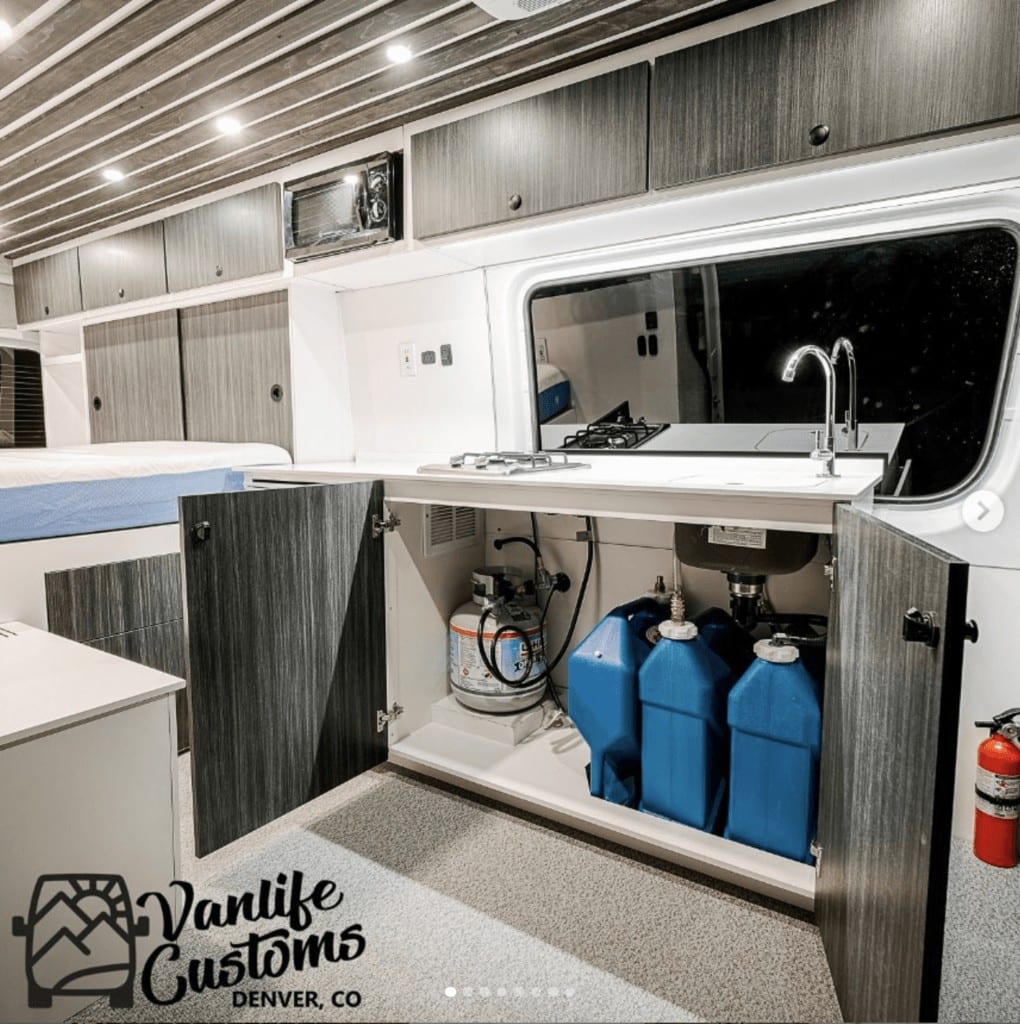 a campervan kitchen with the under-sink doors open, showing 3 5-gallon jugs that are used for the van water system