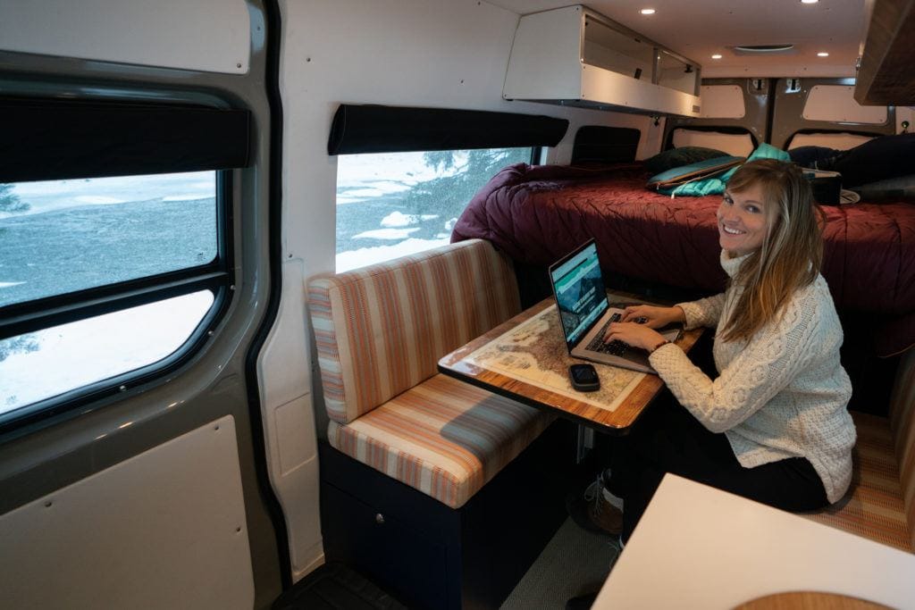 A woman smiles at the camera working from her Sprinter van next to an open laptop.