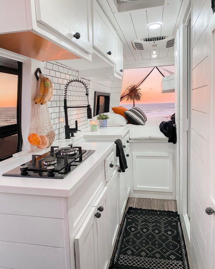 Beautiful campervan kitchen with inlaid propane stove, sink and faucet, and platform bed at rear with doors open to beach sunset