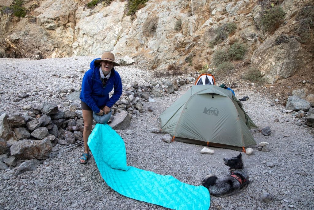 A woman inflates a sleeping pad next to her tent on a backpacking trip