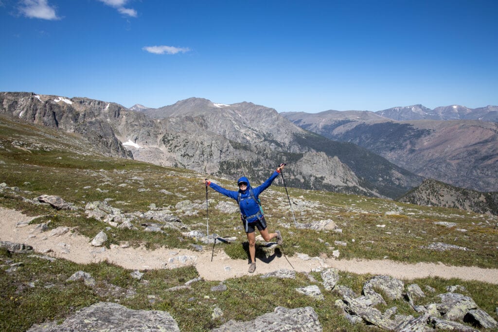 Kristen posing for photo on trail with arms outstretched holding trekking poles and standing on one foot. She is wearing a Patagonia Houdini jacket on a very windy day in Rocky Mountain National Park