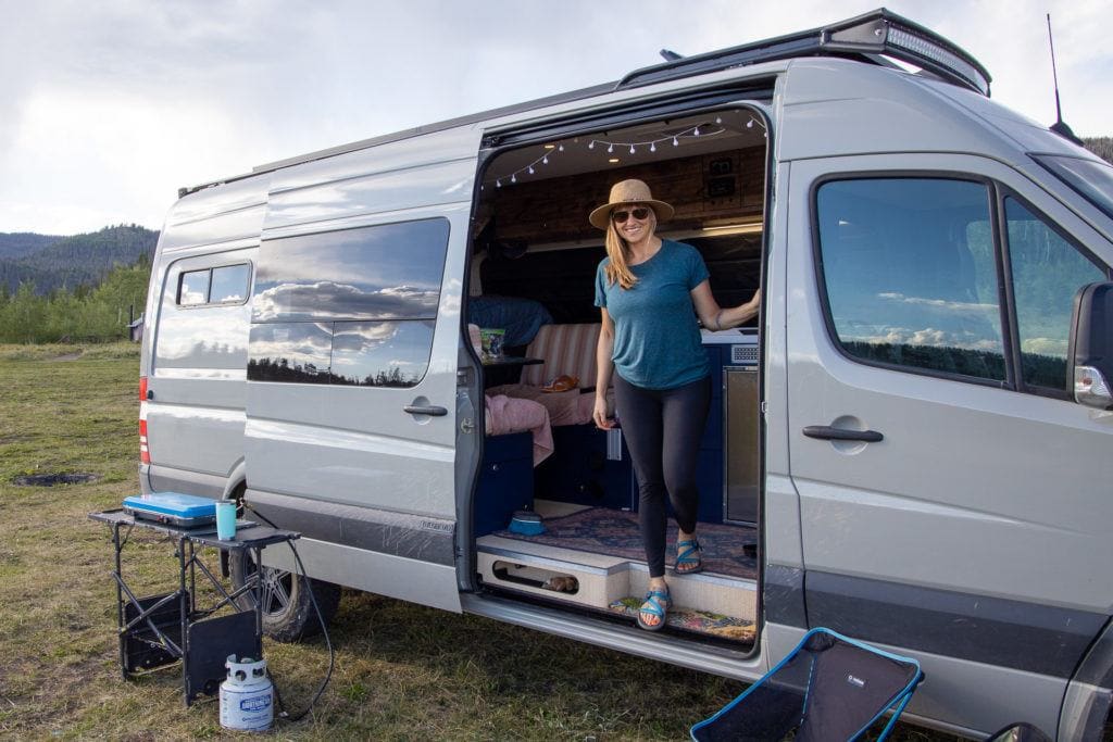 Kristen standing in open doorway of converted camper van at dispersed campsite with cook station set up outside