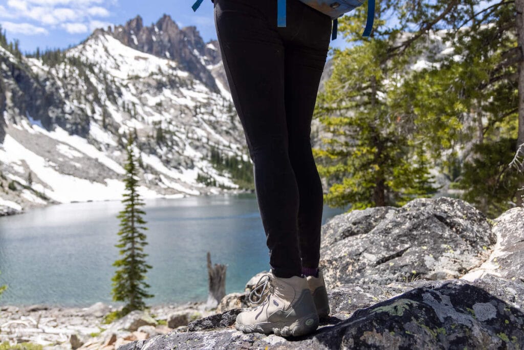 Photo of women's legs and high-ankle hiking boots standing on rock overlooking alpine lake with snow-covered peaks in the background