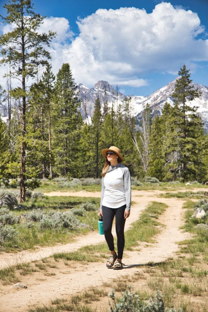 Woman walking on doubletrack dirt road wearing Teva Universal Trail sandals, leggings, long-sleeve shirt, sunglasses, and sun hat