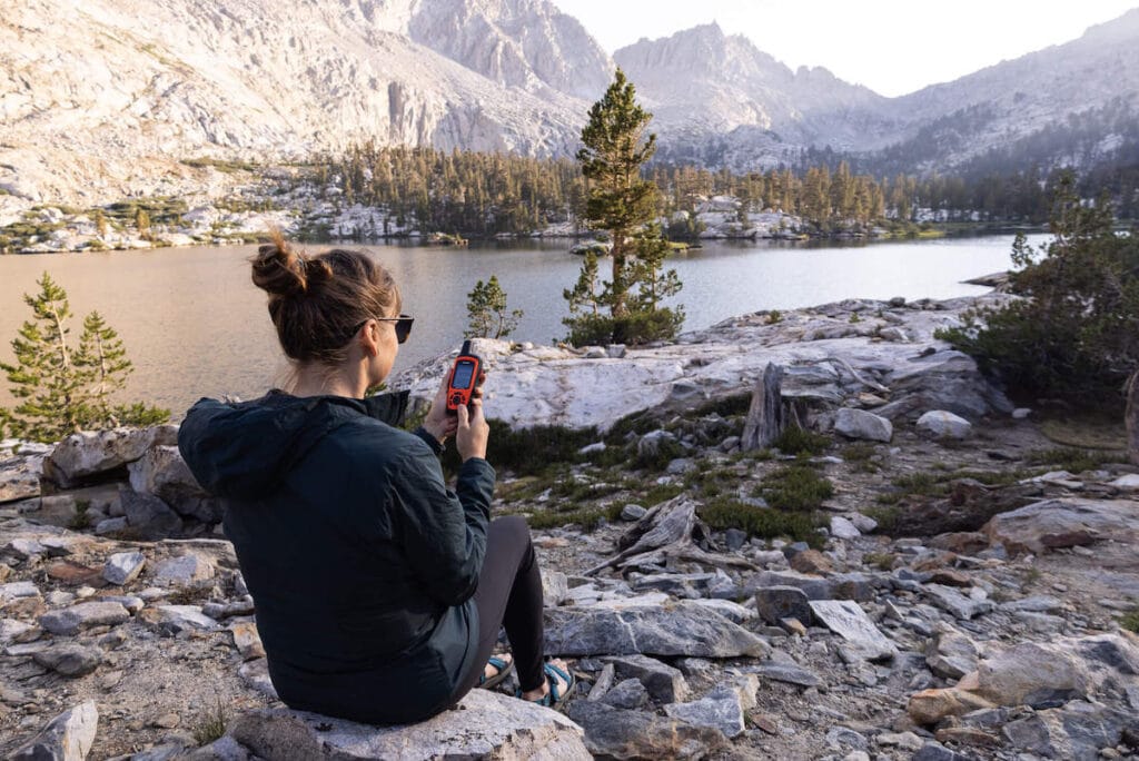 Woman sitting on rock in front of alpine lake and mountain looking at Garmin inReach gps device