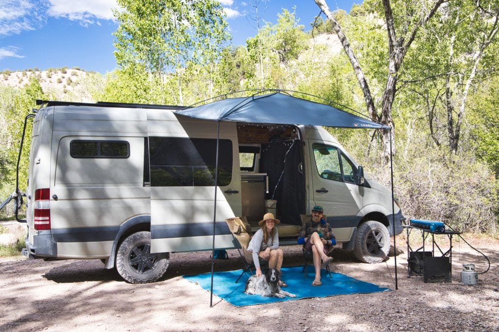 A man and woman sitting in camp chairs under a MoonShade awning attached to camper van roof