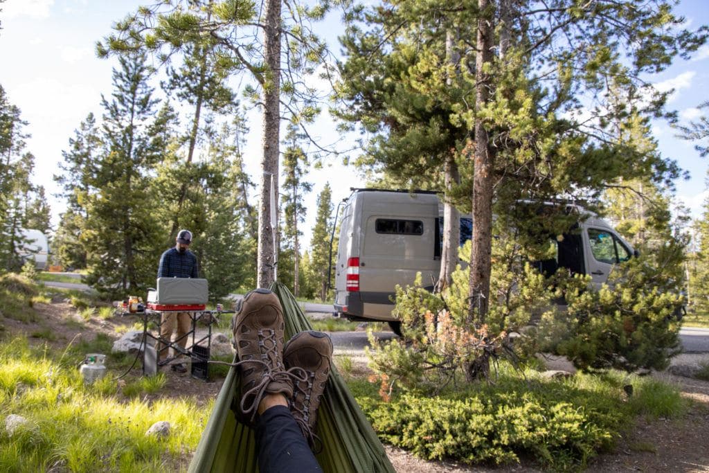 Camper relaxing in hammock at campsite while partner cooks meal on camp stove