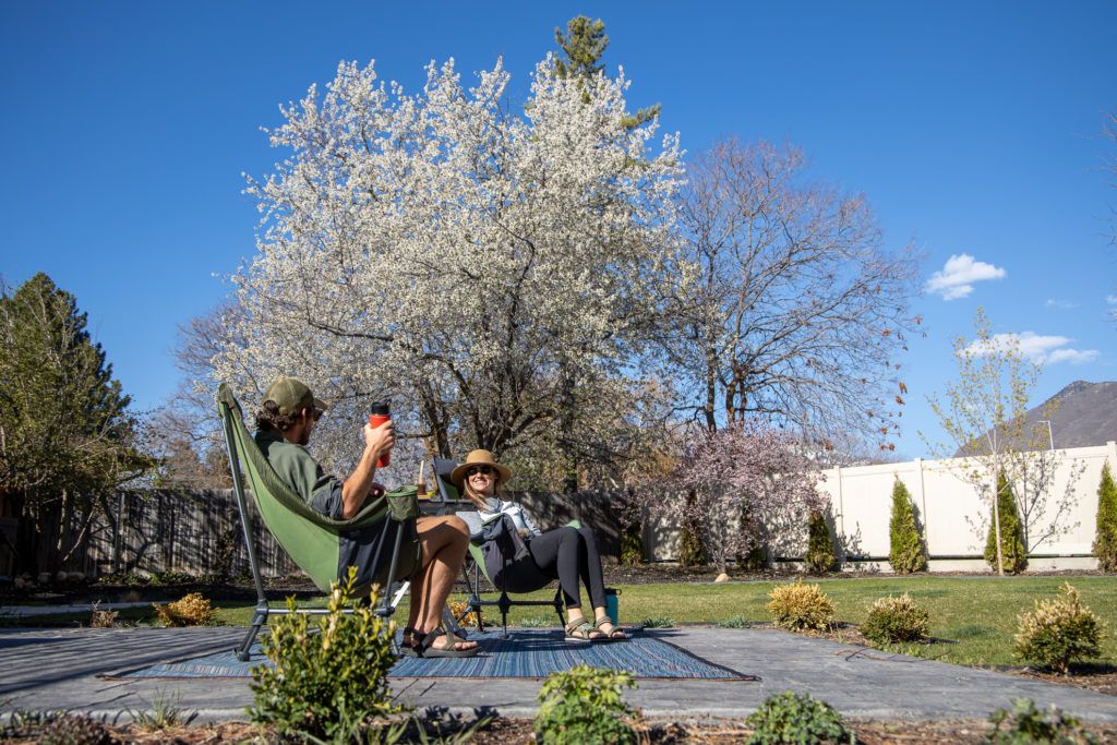 Two people sitting in camp chairs on concrete pad surrounded by trees and green lawn